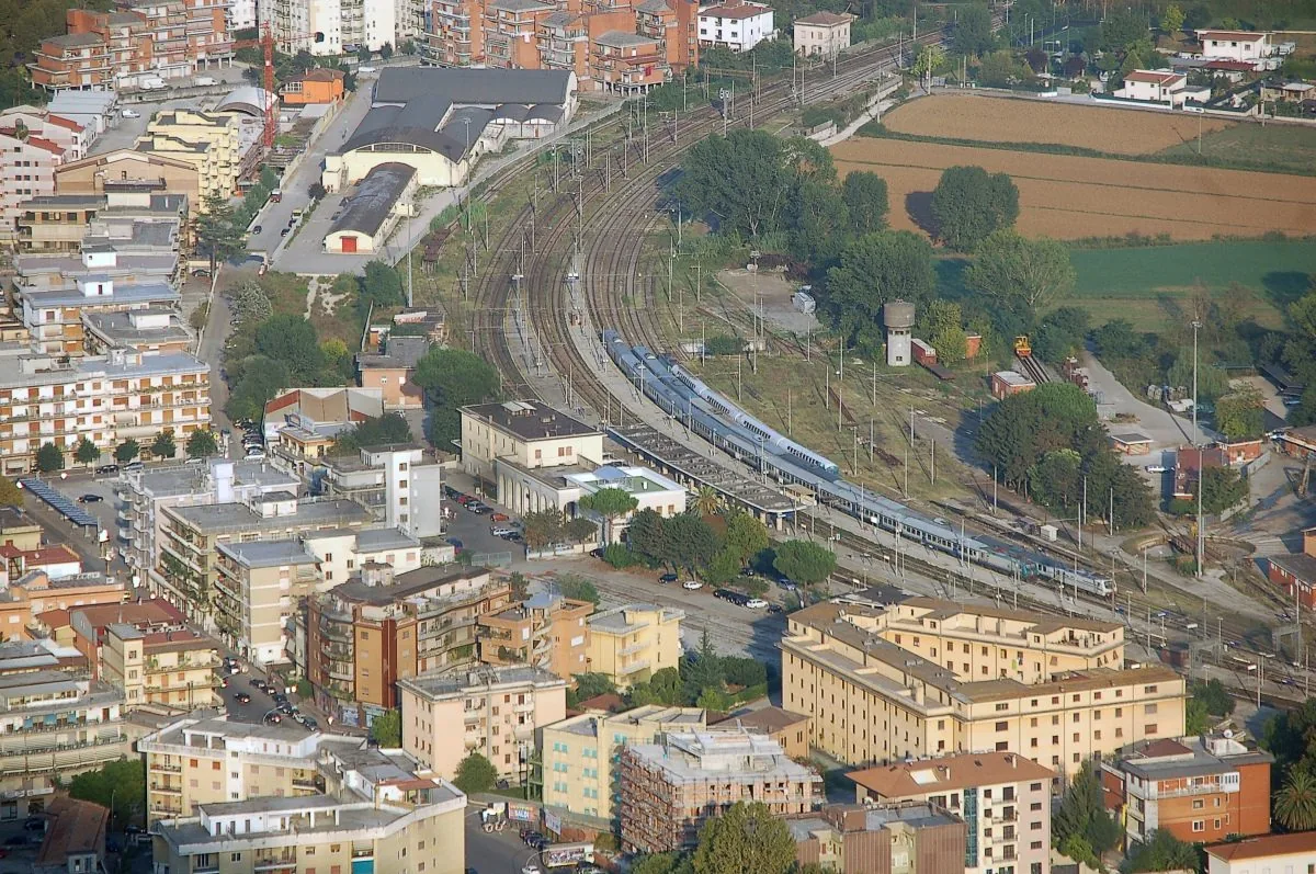 Veduta dall'alto di Cassino con il fascio di binari della stazione ferroviaria e un treno fermo accanto ai marciapiedi, tra palazzi e campi