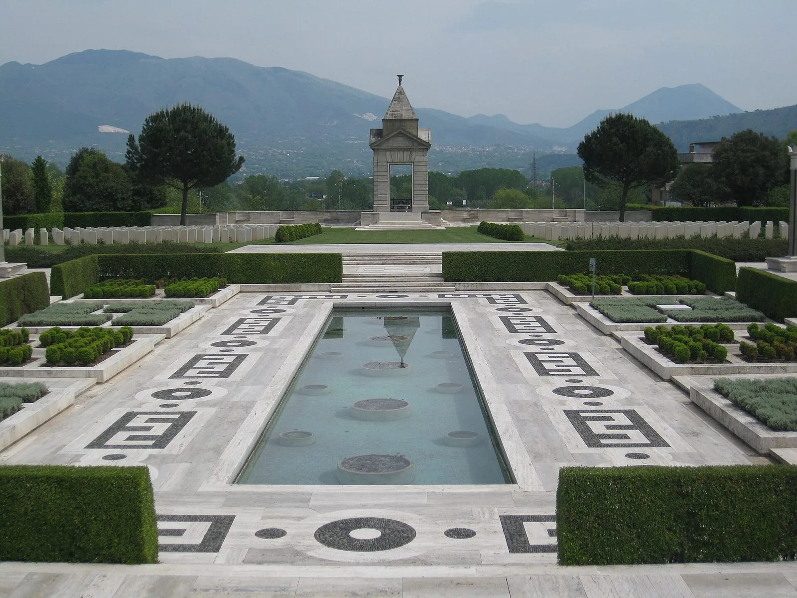 Cimitero militare del Commonwealth a Cassino: vasca d'acqua tra siepi e pavimentazione a motivi geometrici, file di lapidi bianche e memoriale in pietra, montagne sullo sfondo
