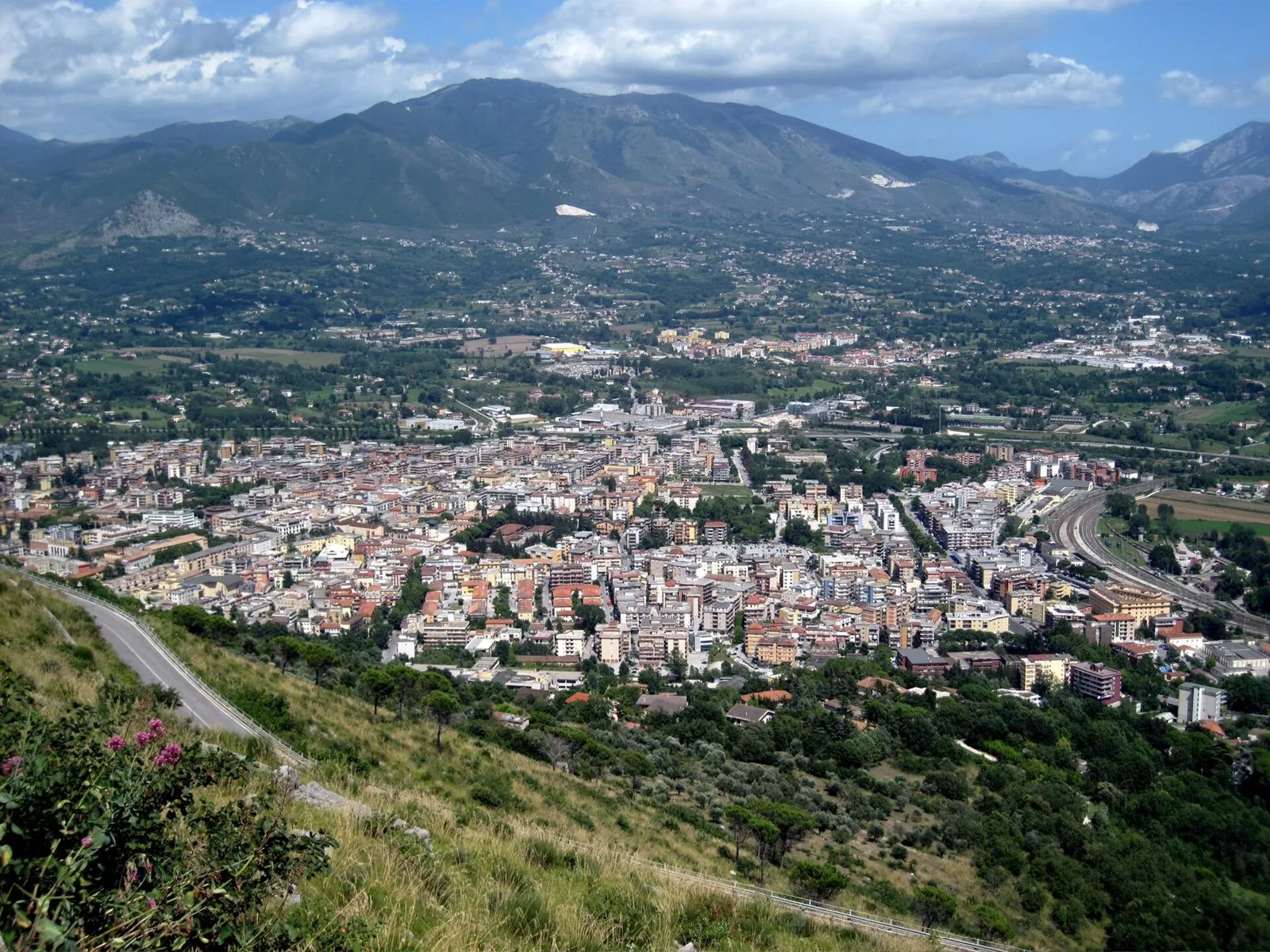 Panorama di Cassino dalla strada che sale verso Montecassino: in primo piano la carreggiata sul pendio, poi la città, la ferrovia e le montagne sullo sfondo